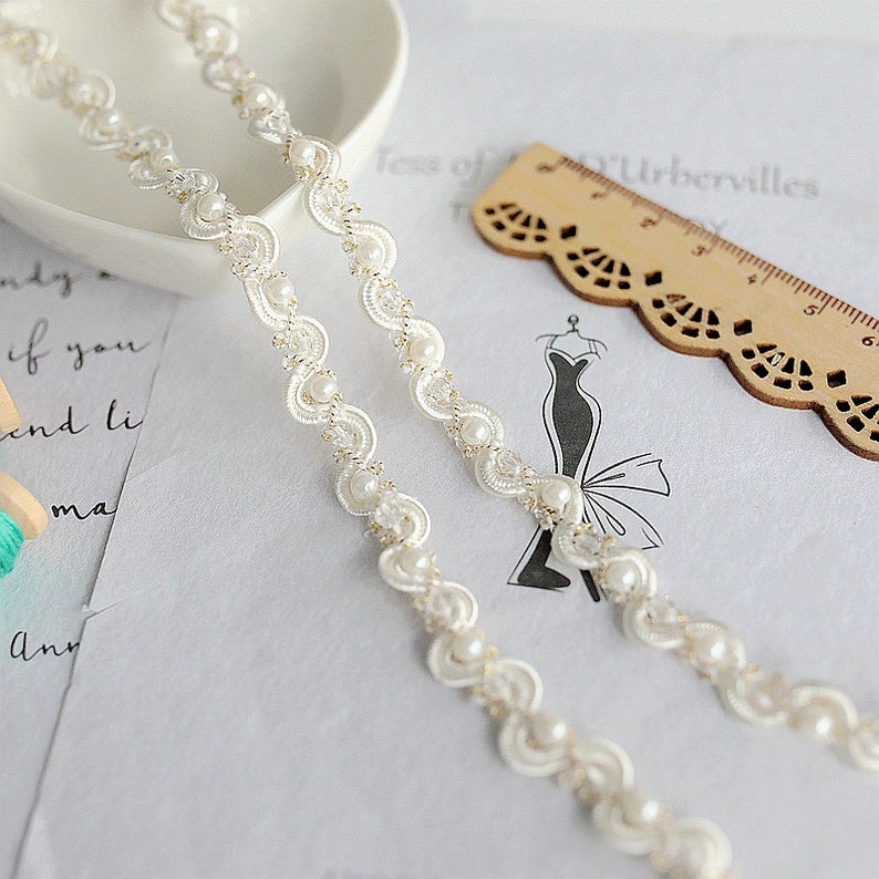 May include: White beaded trim with a scalloped edge. The trim is made of small white pearls and a delicate lace pattern.