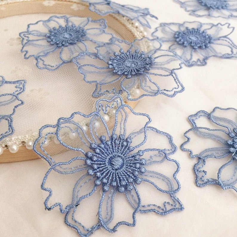 May include: Multiple light blue floral lace appliqu&eacute;s featuring intricate embroidered petals and a textured center. These delicate, sheer fabric flowers are arranged around a circular wooden embroidery hoop frame decorated with small white beads.