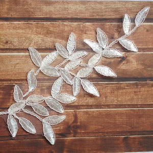May include: A silver leaf lace applique with a delicate floral design. The lace is on a white mesh background and is perfect for adding a touch of elegance to any project.