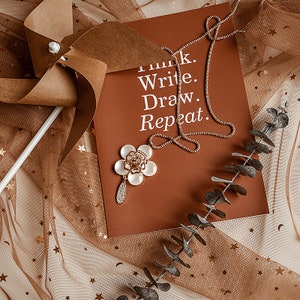 May include: A brown card with the text "Think. Write. Draw. Repeat." on it. A gold chain necklace with a white flower pendant is on top of the card. The card is on a brown and white mesh fabric with gold stars.
