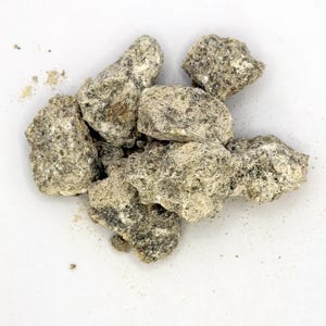 May include: A pile of irregularly shaped, light gray rocks with a speckled texture. The rocks are clustered together on a white surface, with some small particles scattered around them. The overall appearance suggests a natural, earthy material.