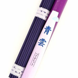 Pode incluir: Um feixe de varetas de incenso roxo escuro presas com faixas de papel, ao lado de um tubo cilíndrico roxo. O tubo apresenta um rótulo branco com texto japonês azul 青雲 e バイオレット, além de um pequeno ícone de gota azul claro.