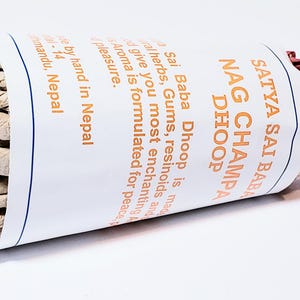 May include: A rolled bundle of Satya Sai Baba Nag Champa Dhoop incense. The white paper wrapping features orange text and a blue border. The incense is made by hand in Nepal and is formulated for peace and pleasure.