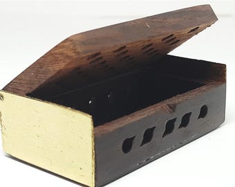 Può includere: Una piccola scatola di legno aperta con coperchio a cerniera. La scatola è marrone scuro con accenti color oro sul davanti e un interno nero. Il coperchio ha intagli decorativi ed è leggermente socchiuso.