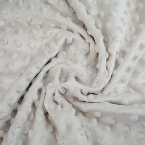 May include: A close-up of a soft, grey, textured fabric with a raised dot pattern. The fabric is wrinkled and folded, showing the texture and depth of the dots.