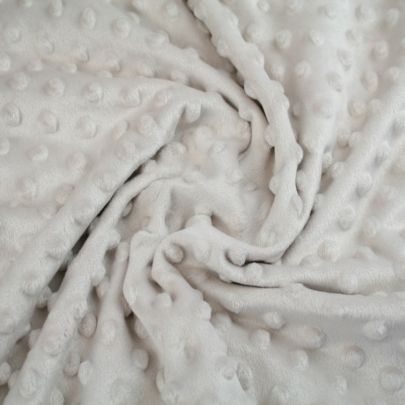 May include: A close-up of a soft, grey, textured fabric with a raised dot pattern. The fabric is wrinkled and folded, showing the texture and depth of the dots.