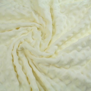 May include: Close-up of a soft, cream-coloured fabric with a textured, bubble-like pattern. The fabric is folded and draped, creating a swirling effect.
