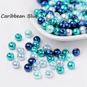 Può includere: Assortimento di perline blu, turchese e bianche dall'aspetto perlato. Le perline sono sparse su una superficie bianca e in una ciotola bianca. Il testo "Caribbean Blue" è visibile nell'immagine.