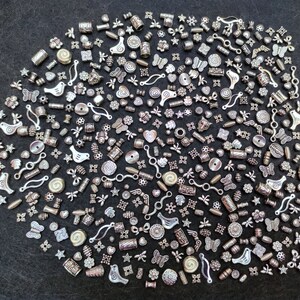 Pu&ograve; includere: Una grande collezione di ciondoli in metallo argentato, tra cui uccelli, farfalle, fiori e forme geometriche. I ciondoli sono disposti in un motivo casuale su uno sfondo grigio scuro.