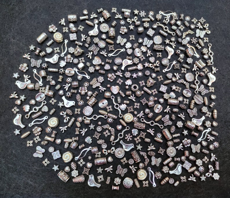Pu&ograve; includere: Una grande collezione di ciondoli in metallo argentato, tra cui uccelli, farfalle, fiori e forme geometriche. I ciondoli sono disposti in un motivo casuale su uno sfondo grigio scuro.