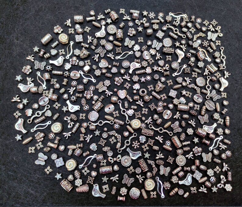Pu&ograve; includere: Una grande collezione di ciondoli in metallo argentato, tra cui farfalle, uccelli, fiori, stelle e forme geometriche. I ciondoli sono sparsi su uno sfondo grigio scuro.