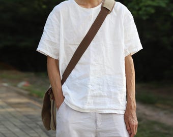 May include: A white linen t-shirt with a crew neck and short sleeves. The shirt is paired with light-coloured trousers. A brown shoulder bag is worn across the body.