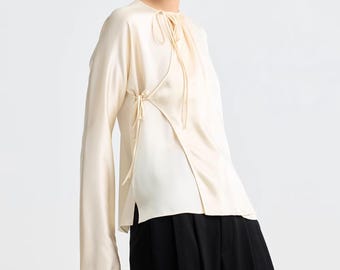 Puede incluir: Blusa crema de manga larga con un diseño asimétrico único. La blusa presenta cierres de lazo en el cuello y en el lateral, y se combina con pantalones negros. La tela tiene un brillo sedoso.
