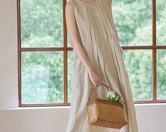 May include: A sleeveless, beige linen dress with pleats at the neckline and a flowing skirt. The dress is paired with a woven basket bag with a handle, filled with white flowers. The background features a window with a view of green foliage.