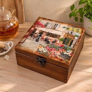 Peut inclure: Une boîte carrée en bois avec un couvercle à charnière orné d'une image imprimée d'une scène de café parisien avec des tables, des chaises et un étal de fleurs. La boîte a une finition brun foncé et un loquet en métal pour fermer le couvercle.