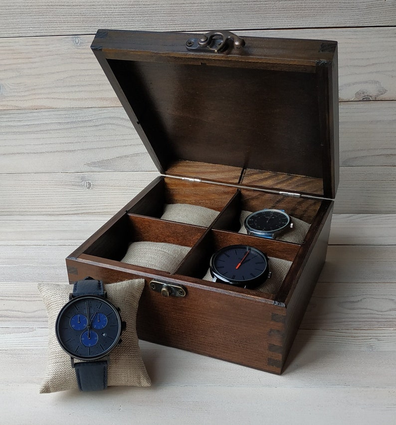 A square wooden watch box with a hinged lid, showing four compartments with beige linen pillows. Three watches rest inside, while one more is on a pillow in front. The dark wood with a metal clasp adds a classic, elegant look.
