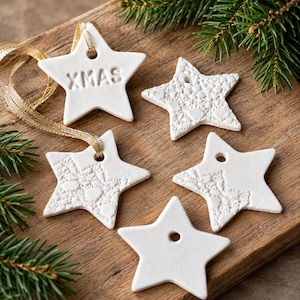 Op de afbeelding: Vijf witte stervormige ornamenten van klei, elk met een gat om op te hangen. Eén ster is voorzien van de tekst XMAS, terwijl andere sterren textuurpatronen of gladde oppervlakken hebben. Een gouden lint is door twee van de sterren geregen.