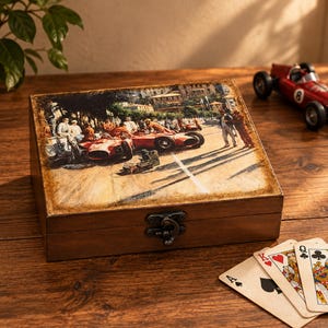 May include: A rectangular wooden box with a hinged lid featuring a printed illustration of vintage red race cars and people at a track. The box has a dark brown finish and a metal latch closure on the front, positioned on a wooden surface.