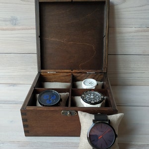 May include: A wooden watch box with a dark brown finish and a hinged lid. The box has six compartments for storing watches. Four watches are visible in the compartments, including a black watch with blue accents, a white watch with black accents, a white watch with black accents, and a black watch with a white face. The box is sitting on a white wooden surface.
