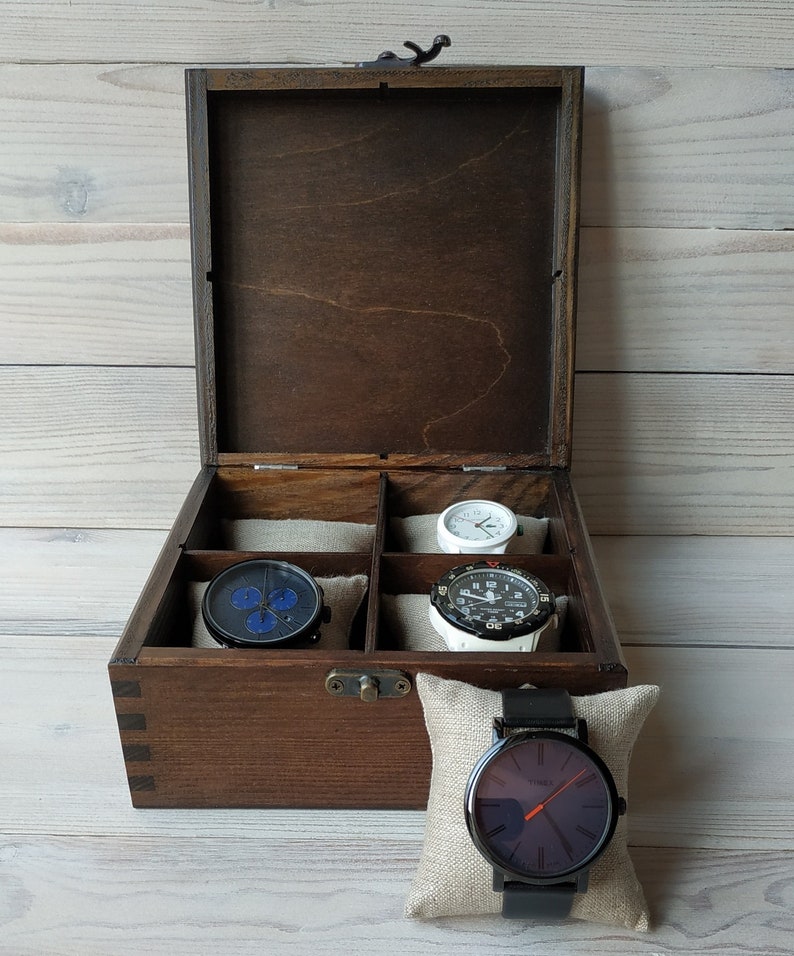 May include: A wooden watch box with a dark brown finish and a hinged lid. The box has six compartments for storing watches. Four watches are visible in the compartments, including a black watch with blue accents, a white watch with black accents, a white watch with black accents, and a black watch with a white face. The box is sitting on a white wooden surface.