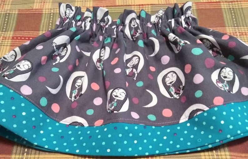 May include: A gathered skirt featuring a dark gray fabric with a repeating pattern of a female character with long dark hair, white crescent moons, and multicolored polka dots, finished with a teal hem band decorated with small white polka dots.