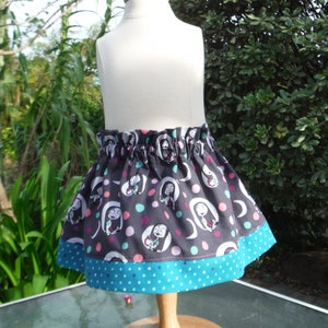 May include: A gathered skirt featuring a dark gray fabric with a repeating pattern of cartoon characters in white ovals, interspersed with pink and teal polka dots. The skirt has a wide teal hem with white polka dots and an elasticized waistband.