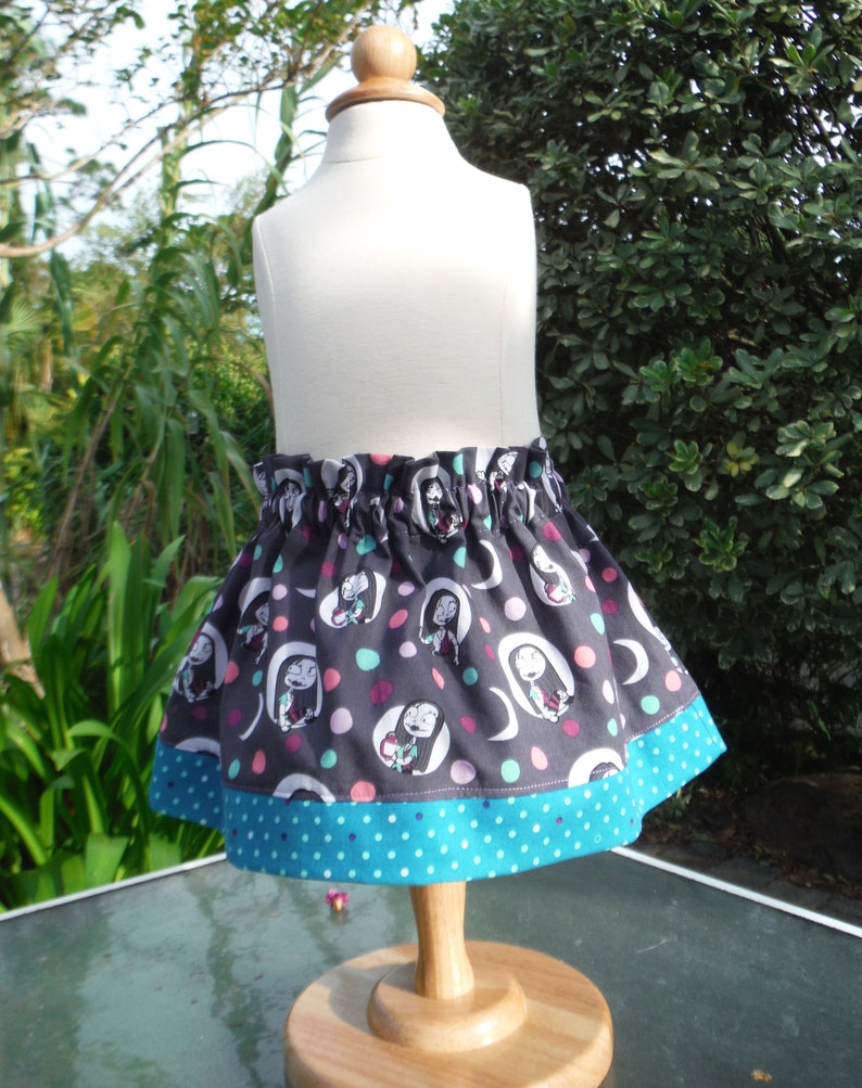 May include: A gathered skirt featuring a dark gray fabric with a repeating pattern of cartoon characters in white ovals, interspersed with pink and teal polka dots. The skirt has a wide teal hem with white polka dots and an elasticized waistband.