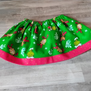 May include: A green skirt with a red trim featuring cartoon characters and snowflakes. The skirt has an elastic waistband and is decorated with images of a Grinch-like character, gifts, and snowflakes. The trim is a solid red color.