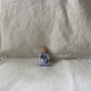 Könnte beinhalten: Eine kleine, handgefertigte Puppe mit blonden Haaren, die ein blau-weiß gestreiftes Kleid trägt. Die Puppe sitzt auf einer weißen Stoffoberfläche.