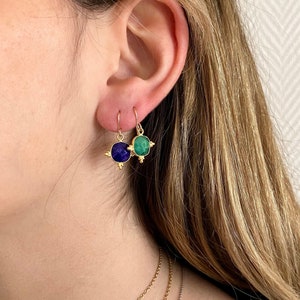 Puede incluir: Pendientes dorados con una piedra cuadrada azul oscuro y una piedra cuadrada verde. Los pendientes tienen un pequeño diseño decorativo en cada esquina. Se muestran en la oreja de una persona.