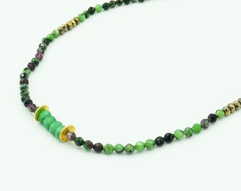 May include: A delicate necklace featuring small, faceted beads in shades of green, black, and burgundy. The necklace includes gold-colored accents and a cluster of green beads with gold-colored discs. The necklace is on a white background.