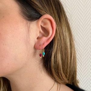 Puede incluir: Pendientes colgantes dorados con piedras preciosas en forma de lágrima. Un pendiente presenta una piedra roja, el otro una turquesa. Los pendientes cuelgan de un gancho simple y tienen pequeños detalles dorados.