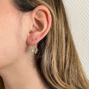 Puede incluir: Pendientes dorados con piedras preciosas rectangulares de colores. Un pendiente presenta una piedra verde azulado claro, el otro una piedra marrón claro. Los pendientes cuelgan de delicados ganchos dorados y tienen pequeños detalles dorados.