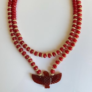 Peut inclure: Un collier à plusieurs rangs avec des perles rouges et des entretoises dorées. Le collier comporte un pendentif rouge sculpté en forme d'aigle. Le collier est présenté sur fond blanc. Bijoux.