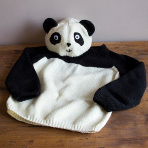 May include: A white and black knitted sweater with a panda bear hood. The sweater has long sleeves and a black band around the neck.