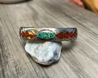 May include: A silver cuff bracelet with inlaid turquoise and coral-colored stones. The bracelet features a central oval of turquoise stones, flanked by red arrow-shaped designs. The metal has a detailed, etched pattern.