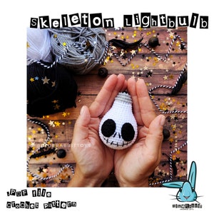 Puede incluir: Una bombilla de crochet blanca con un diseño de calavera y huesos cruzados negros. La bombilla está siendo sostenida en las manos de una persona. El fondo es una superficie de madera con estrellas negras y doradas.
