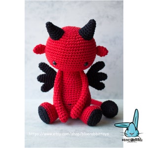 Puede incluir: Un juguete de peluche de diablo rojo tejido a crochet con cuernos, alas y pies negros. El juguete est&aacute; sentado sobre una superficie blanca. El juguete tiene una direcci&oacute;n de sitio web en la parte inferior: https://www.etsy.com/shop/bluerabbittoys