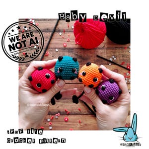 May include: Four small, round, crocheted amigurumi figures in red, teal, orange, and purple, each featuring black horns and large black eyes. The text Baby devil and PDF file crochet pattern is visible along with a Blue Rabbit logo.