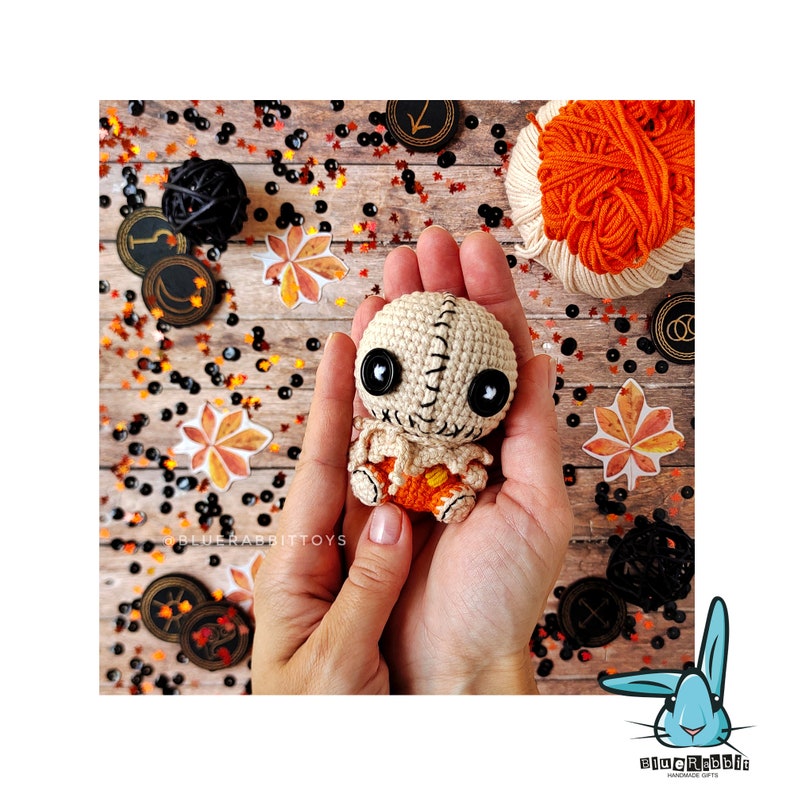 May include: A crocheted doll with a stitched-on face, black button eyes, and orange and white clothing. The doll is being held in a person's hand against a background of wooden planks and colorful confetti.