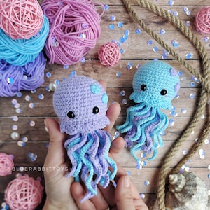 May include: Two crocheted jellyfish toys featuring round heads and curly tentacles. One jellyfish is light purple with light blue spots, and the other is light blue with light purple spots. Both have black eyes and pink cheeks. Text reads @BLUERABBITTOYS.