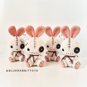 May include: Three crocheted plush bunny toys with pink ears and white bodies. Each bunny has black button eyes, a stitched mouth, and brown yarn accents. The bunnies are sitting on a white surface.