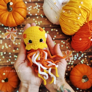 May include: A crocheted jellyfish toy with a yellow head featuring orange spots, black safety eyes, and pink blush marks. The jellyfish has long, dangling tentacles in white, orange, and yellow yarn. The text BLUERABBITTOYS is visible.