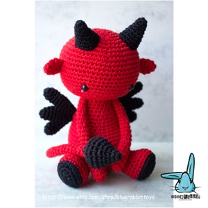 Puede incluir: Un juguete de peluche de diablo rojo tejido a crochet con cuernos, alas y cola negros. El juguete est&aacute; sentado con las piernas cruzadas y la cola enroscada alrededor de sus pies. El juguete tiene ojos negros y una peque&ntilde;a nariz rosa.