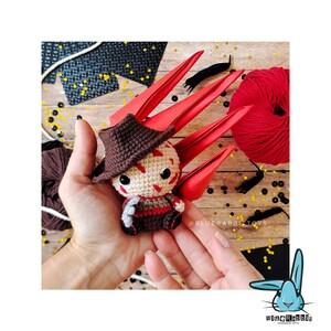 Peut inclure: Un jouet en crochet amigurumi repr&eacute;sentant un personnage de film d'horreur avec un sch&eacute;ma de couleurs rouge et marron. Le jouet tient un couteau et a un sourire effrayant. Le jouet est assis dans une main avec un fond en papier rouge.