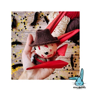 Peut inclure: Un jouet amigurumi en crochet de Freddy Krueger du film A Nightmare on Elm Street. Le jouet porte un chapeau brun et une chemise ray&eacute;e rouge et brune. Le jouet tient un couteau dans sa main droite. Le jouet est assis sur une main avec un fond rouge et blanc.
