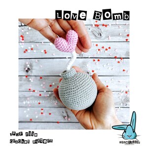 Puede incluir: Un juguete de bomba de ganchillo gris de forma esf&eacute;rica con una mecha blanca rematada por un coraz&oacute;n rosa. El texto "LOVE BOMB" aparece en la parte superior y "PDF file crochet pattern" abajo a la izquierda. Confeti rojo y blanco rodea el objeto.