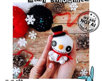 May include: A small white crocheted snowman toy with black button eyes, an orange nose, and a stitched mouth. It wears a black top hat with a red band and a red scarf. The text reads MINI SNOWMAN, WE ARE NOT AI, and .PDF file crochet pattern.