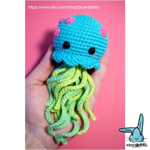 May include: A small, handmade crochet jellyfish toy featuring a bright turquoise rounded head with pink polka dots, two black plastic safety eyes, and long, dangling tentacles in varying shades of light green and yellow yarn.