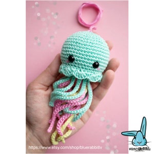May include: A small, crocheted jellyfish toy featuring a mint green rounded head with black plastic eyes and pink stitched cheeks. The jellyfish has multiple dangling, curly tentacles in pastel shades of mint green, pink, and pale yellow.